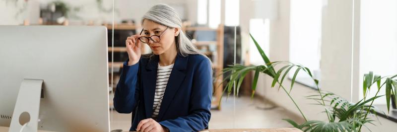 The Asian woman sitting at a table in front of the monitor adjusting glasses with her hand in a light office