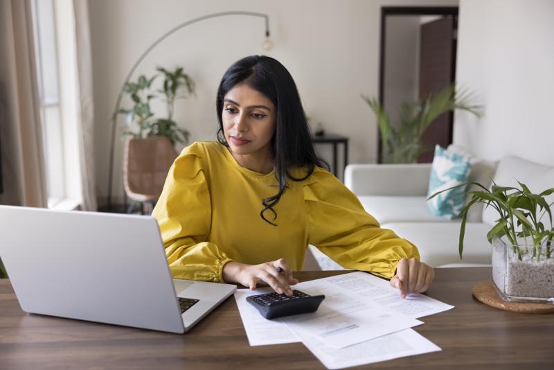 Young Indian woman sit at table with papers and laptop, controlling personal incomes and monthly expenses, reviewing invoices or bills to pay, calculating and preparing tax returns, managing finances
