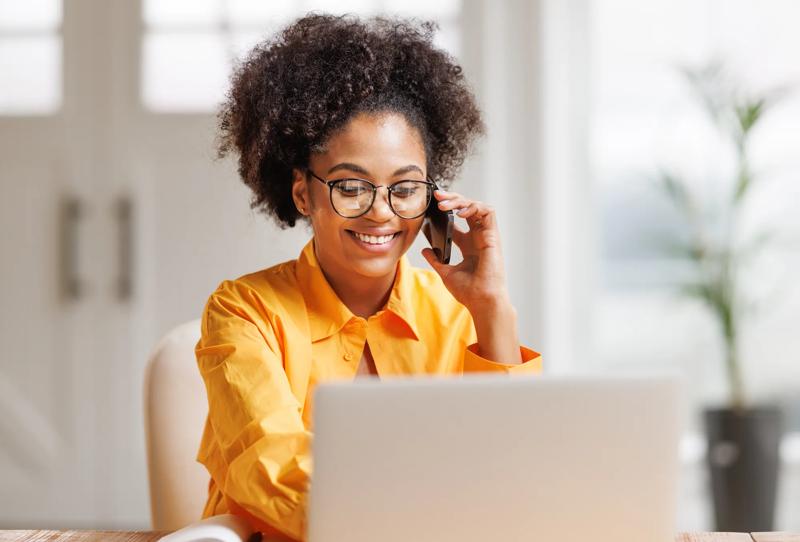 Beautiful smiling african american woman making call via smartphone while working remotely from home while sitting at desk and talking to coworkers.   Ethnic female freelancer talking on mobile phone during remote work