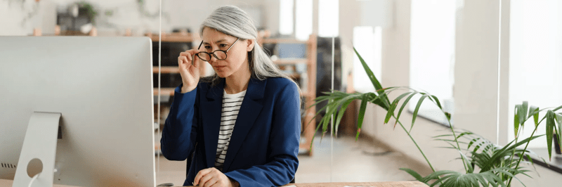 The Asian woman sitting at a table in front of the monitor adjusting glasses with her hand in a light office