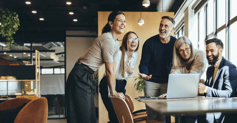 Happy businesspeople laughing while collaborating on a new project in an office. Group of diverse businesspeople using a laptop while working together in a modern workspace.