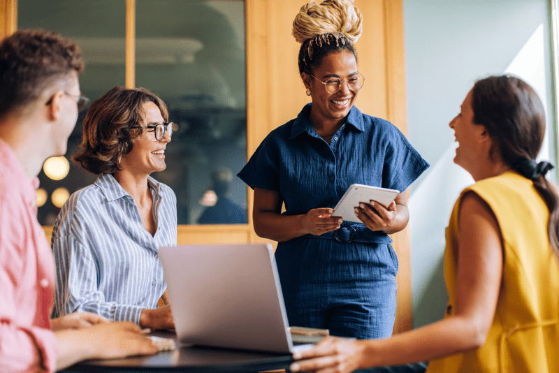 A team of colleagues gather in a modern office, sharing ideas easily. They use laptops and tablets while engaging in a positive and friendly environment, signifying teamwork and creativity.