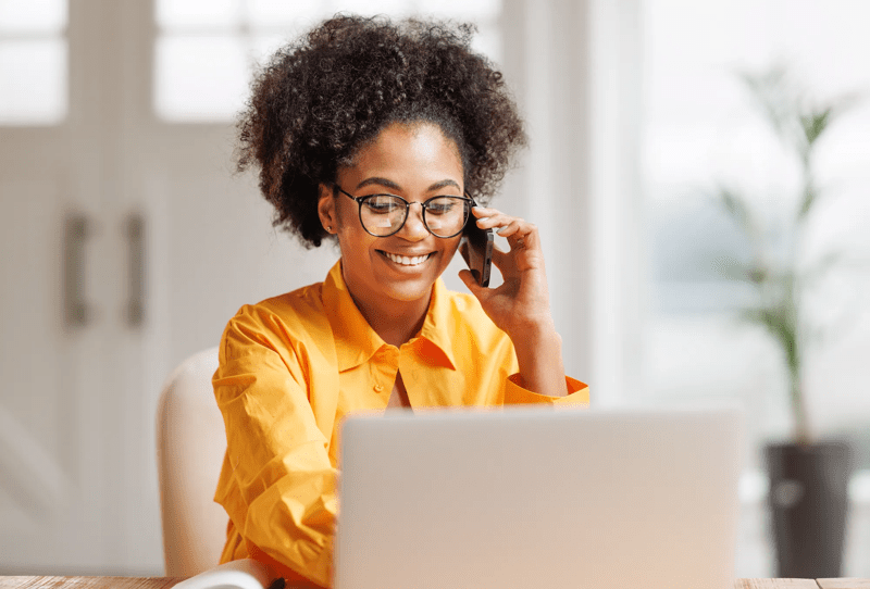 Beautiful smiling african american woman making call via smartphone while working remotely from home while sitting at desk and talking to coworkers.   Ethnic female freelancer talking on mobile phone during remote work