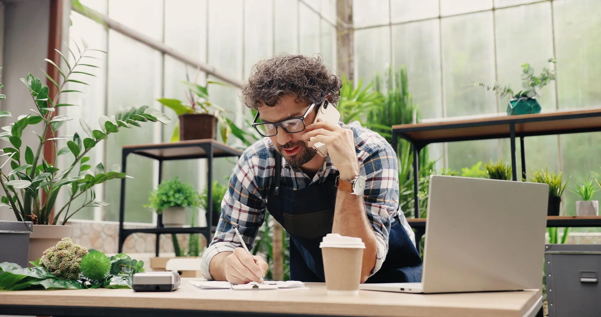 Happy Caucasian businessman talking on cellphone while standing in apron in small floral center and writing down order details. Joyful male florist calling on smartphone at work. Own business concept