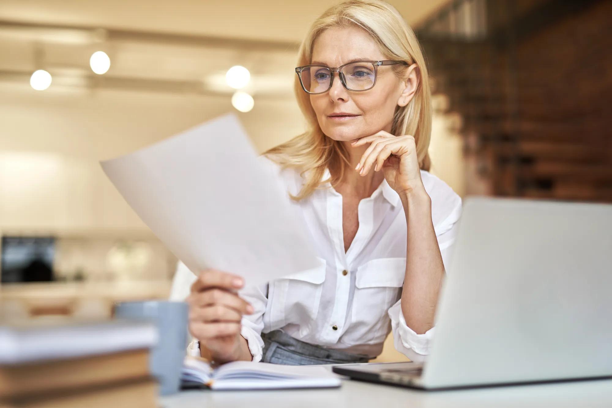 Beautiful mature businesswoman reading papers, using laptop while sitting at her desk in the office. People, business, career concept
