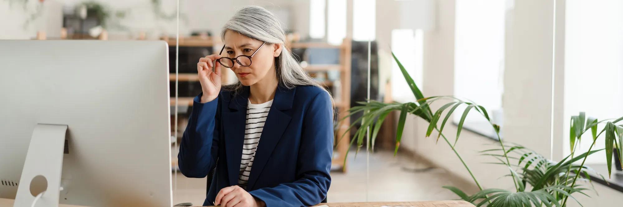 The Asian woman sitting at a table in front of the monitor adjusting glasses with her hand in a light office
