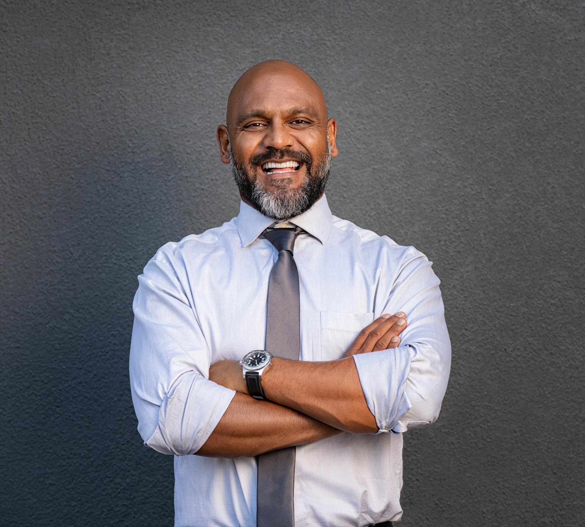 Successful senior man with folded arms standing over grey background. Handsome mature black businessman in shirt and tie looking at camera. Portrait of joyful business man on a grey wall with copy space.