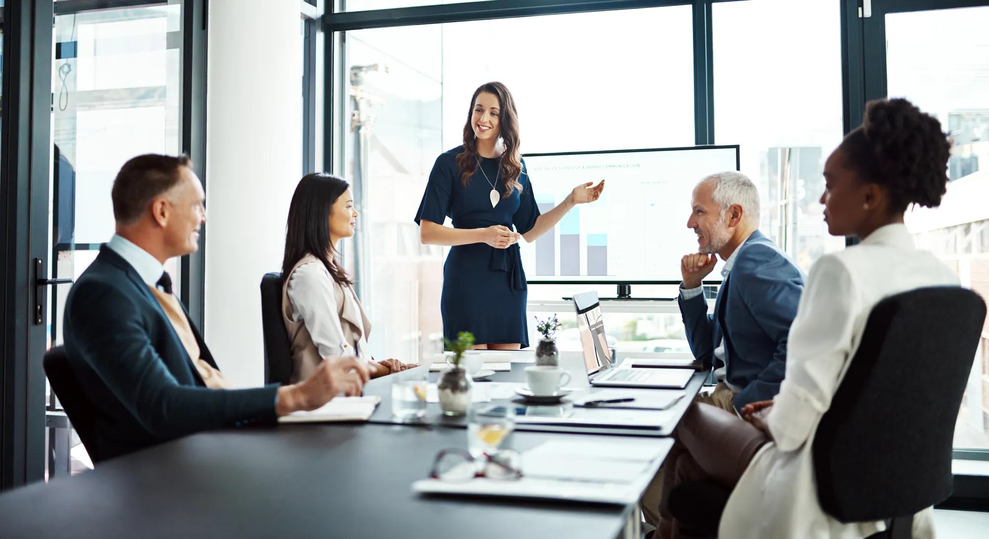 Business woman speaker in an investment meeting with financial data on a boardroom screen. International executive team listening to a presentation. Finance worker talking at a partnership proposal.