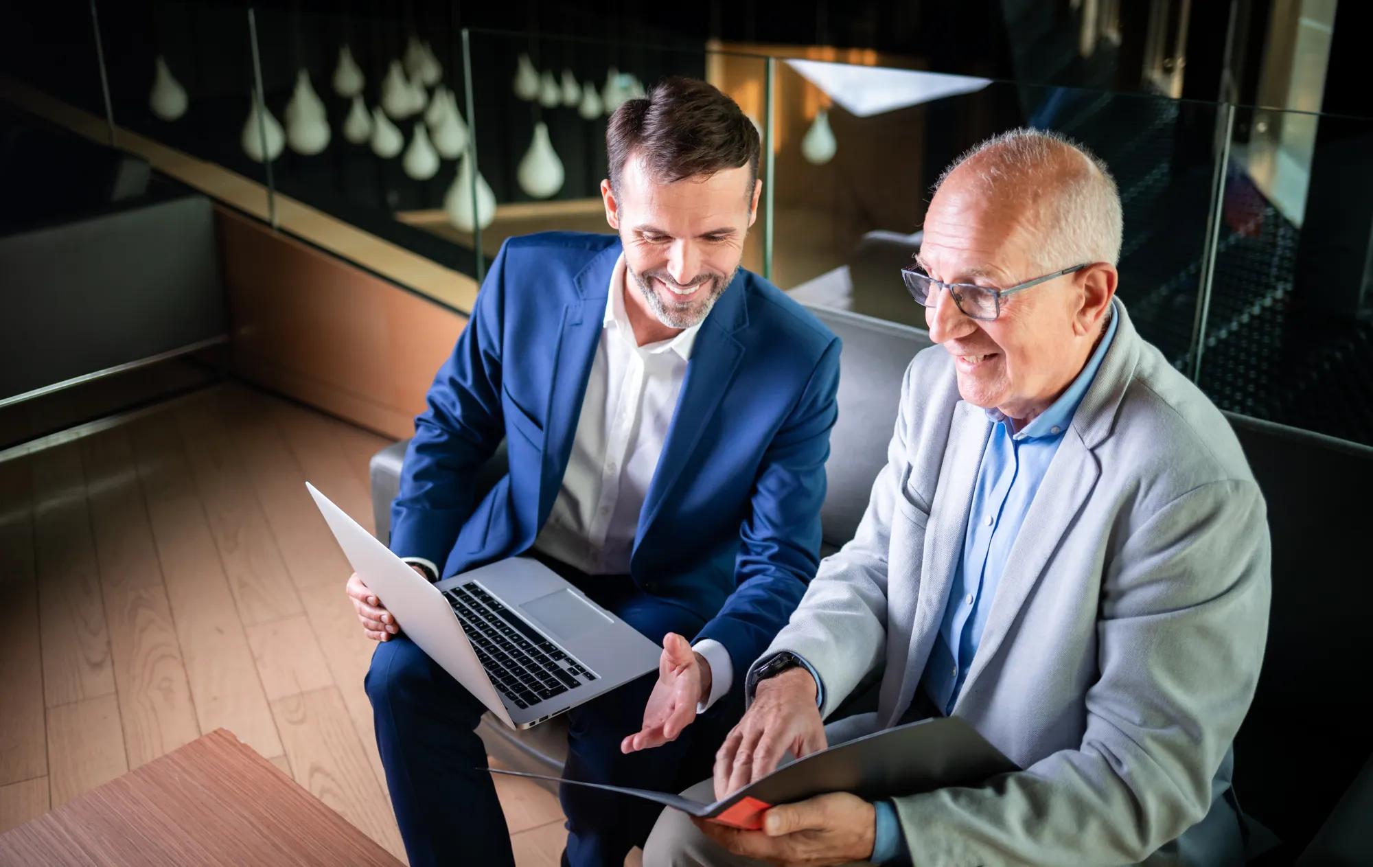 Businessman using laptop to discuss information with older colleague in modern business lounge