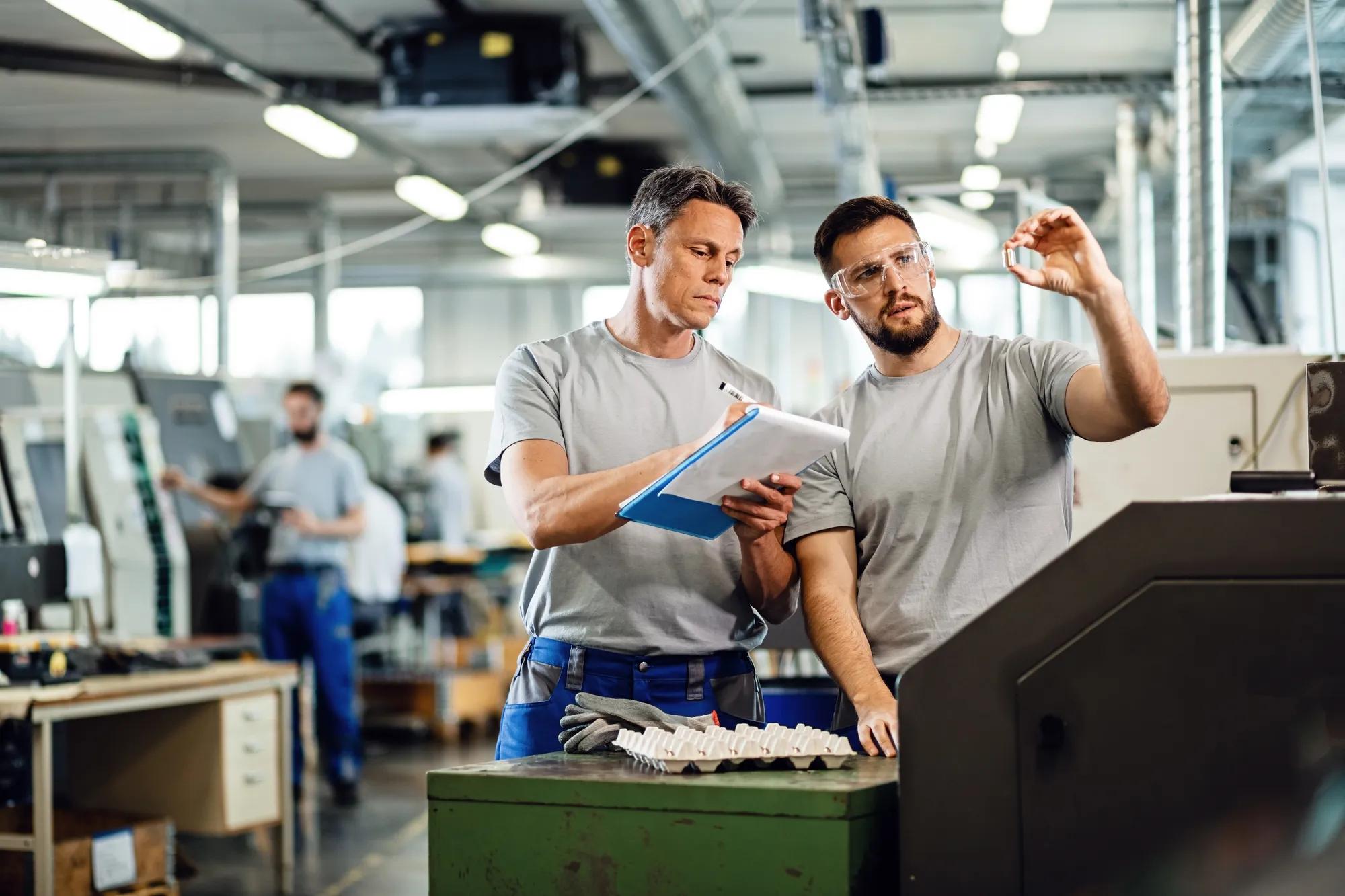 TWO CNC machine operators examining finished products and writing notes in industrial facility. 