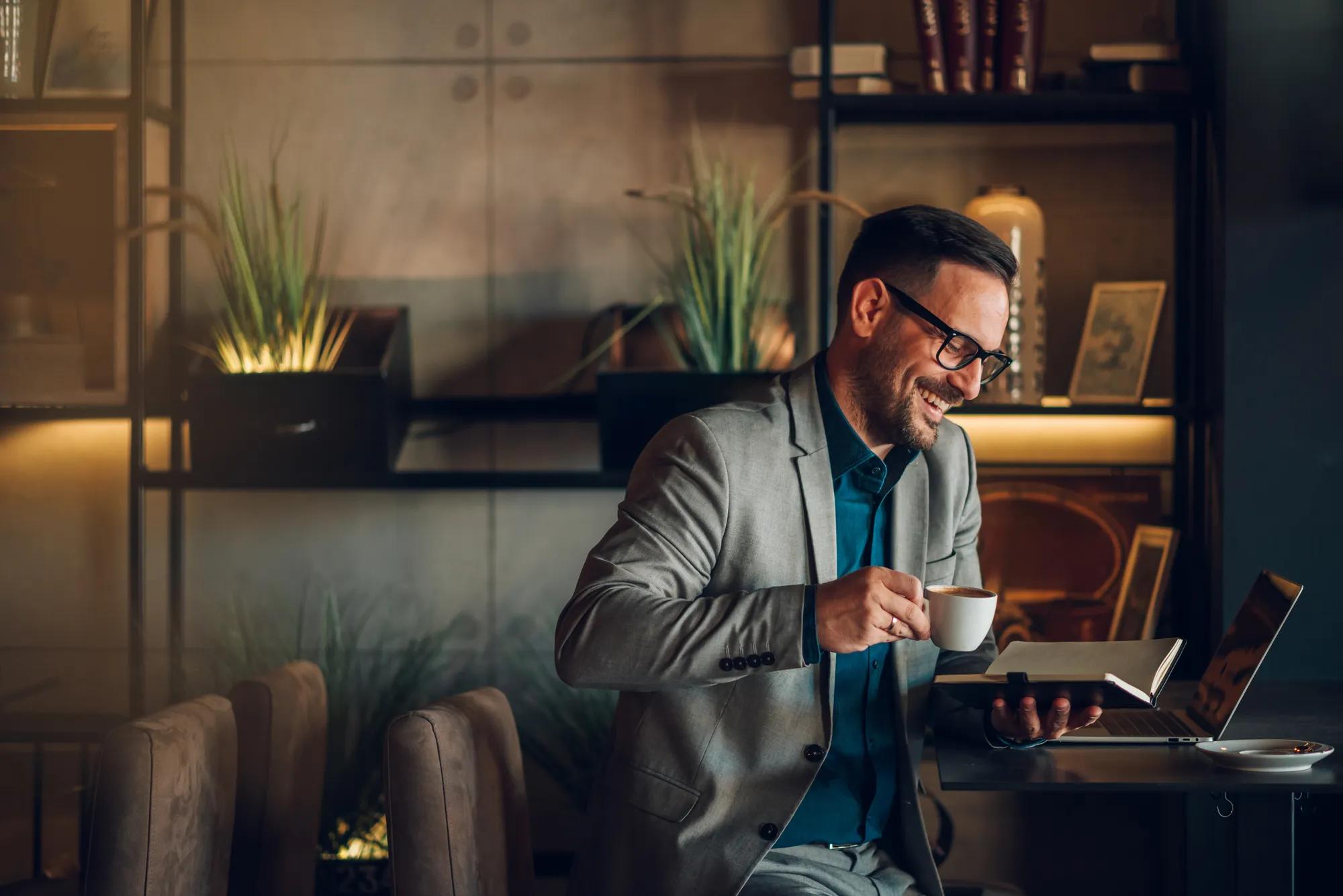 Smiling businessman enjoying coffee while working on a laptop in a stylish modern cafe, embodying productivity and relaxation