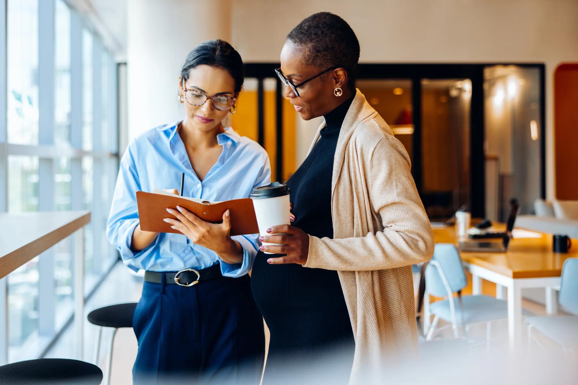 Two businesswomen standing in an office, one holding a clipboard while discussing work and ideas. The other woman is pregnant and holding a beverage cup.