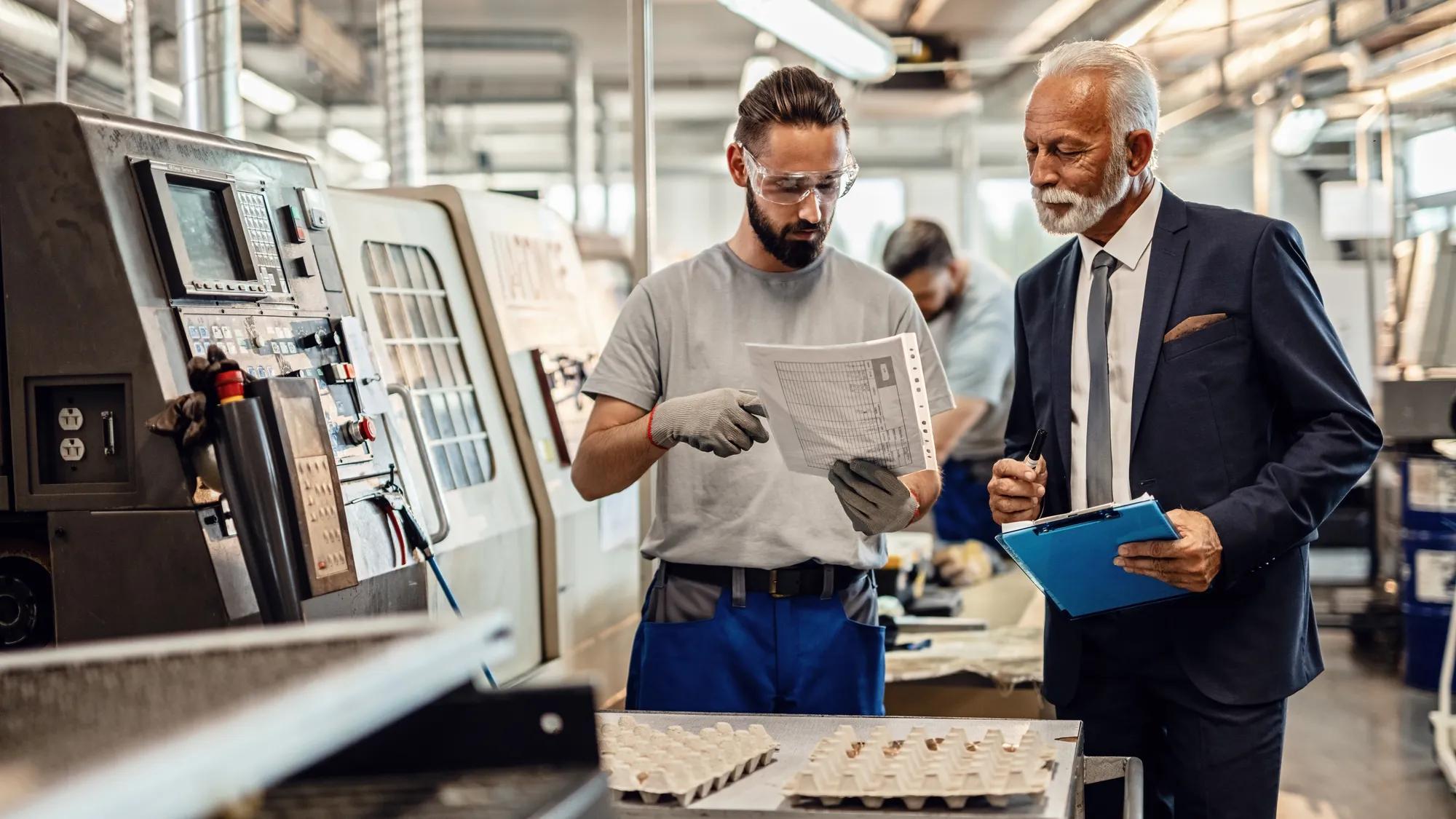 Manual worker and senior businessman cooperating while going through paperwork in factory plant. 