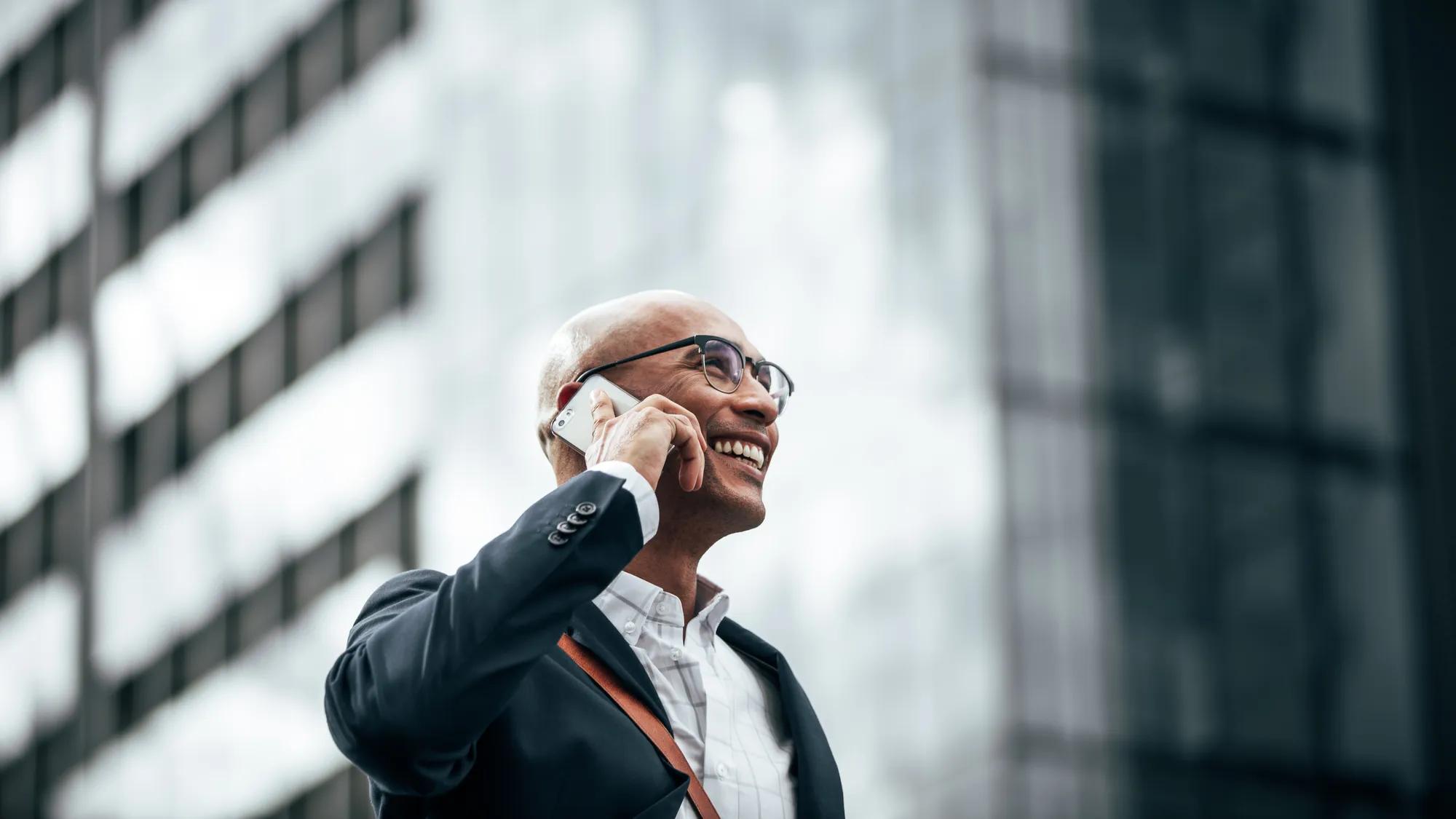 Man in formal clothes wearing office bag walking on street while talking on mobile phone. Smiling businessman talking over cell phone while commuting to office with a glass facade building in background.
