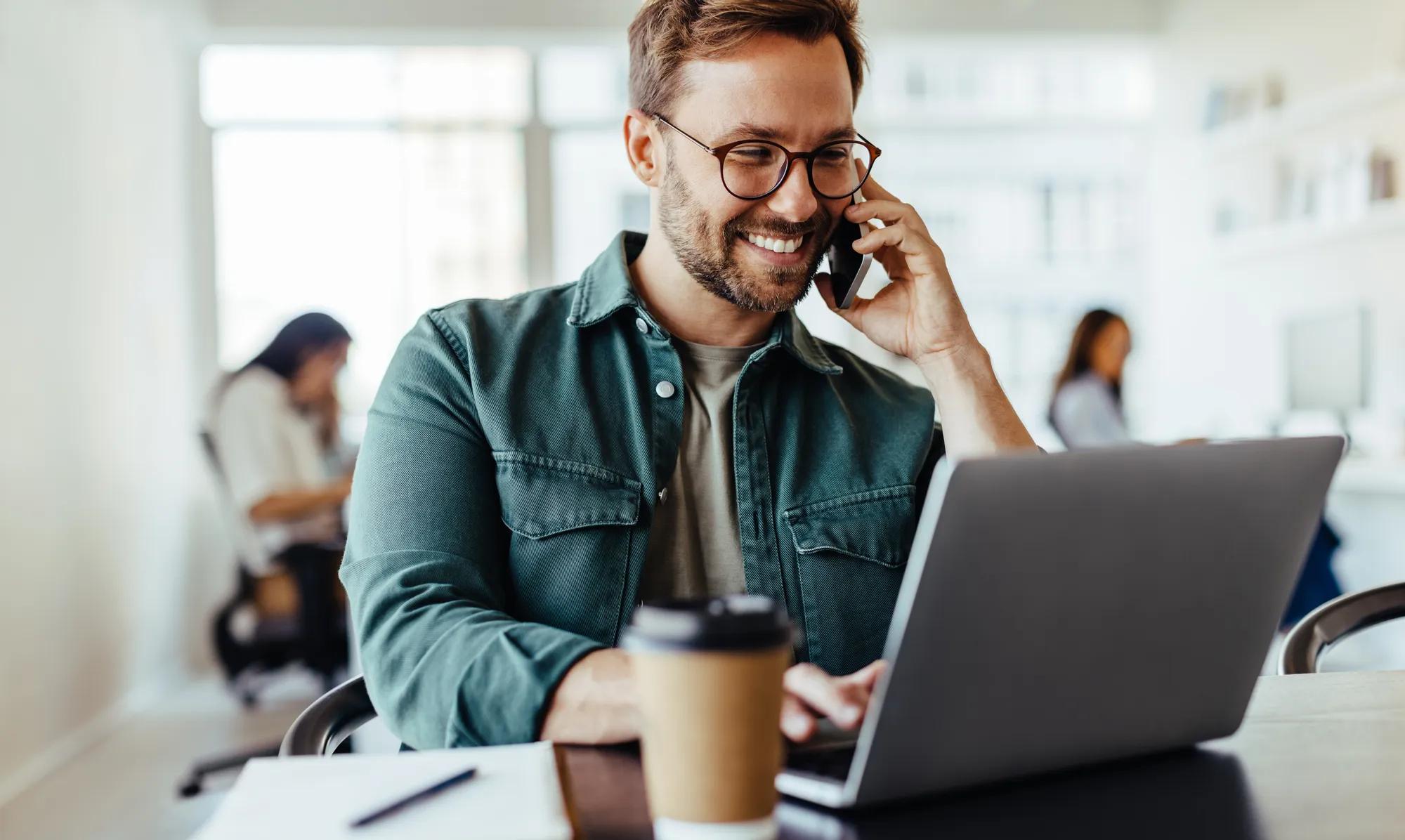 Male software designer speaking to a client on the phone while using a laptop. Happy young business man working in a co-working office.