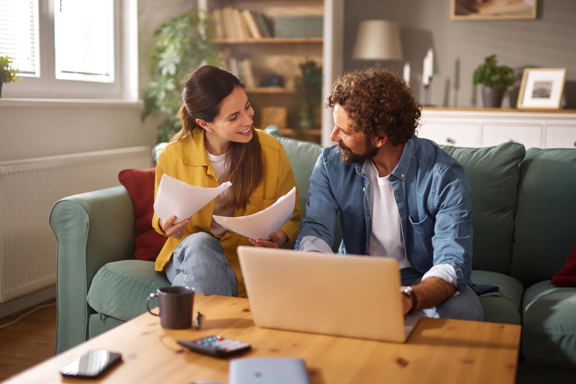 A couple sits together on their couch discussing various life insurance plans. They review important documents while a laptop is open on the coffee table, creating a warm atmosphere.