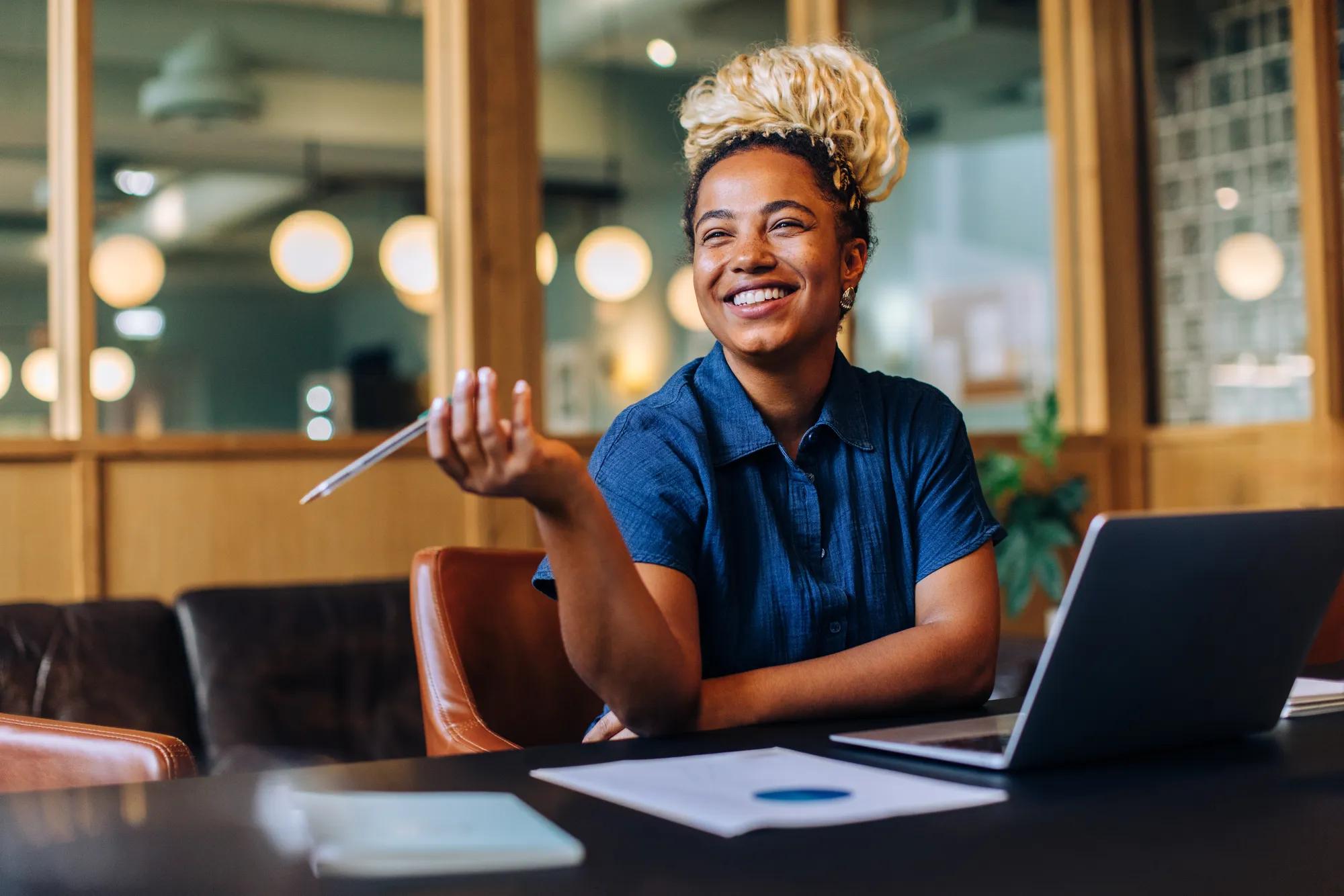 A cheerful young woman is seated at an office desk, holding a pen and smiling openly. She is working with her laptop open, appearing engaged and content in her workplace.