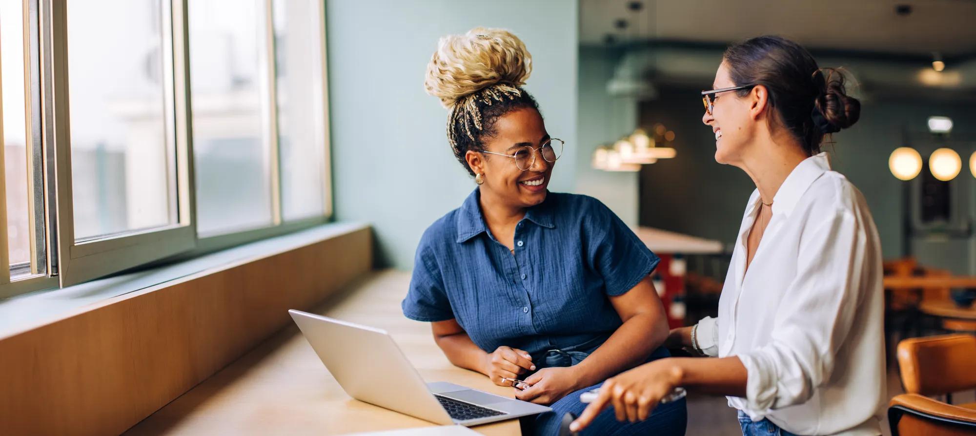 Two women are seated in a bright indoor setting, engaging in conversation near a laptop. They appear to be sharing ideas and enjoying their discussion while wearing casual attire.