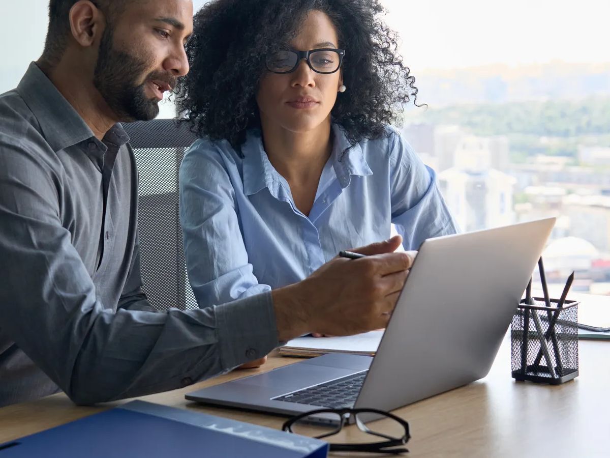 Indian male ceo executive manager mentor giving consultation on financial operations to female African American colleague intern using laptop sitting in modern office near panoramic window.