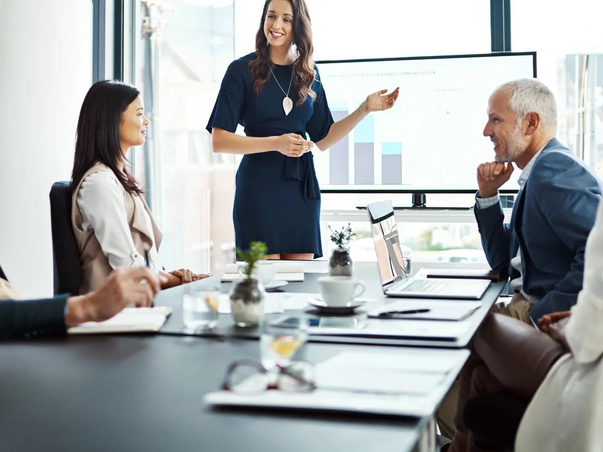 Business woman speaker in an investment meeting with financial data on a boardroom screen. International executive team listening to a presentation. Finance worker talking at a partnership proposal.