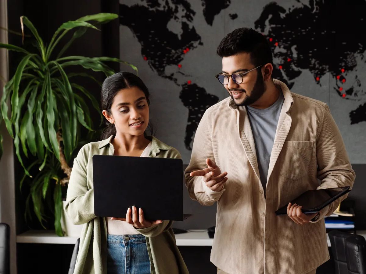 Two young busy indian businesspeople using laptop and tablet while working together in modern office