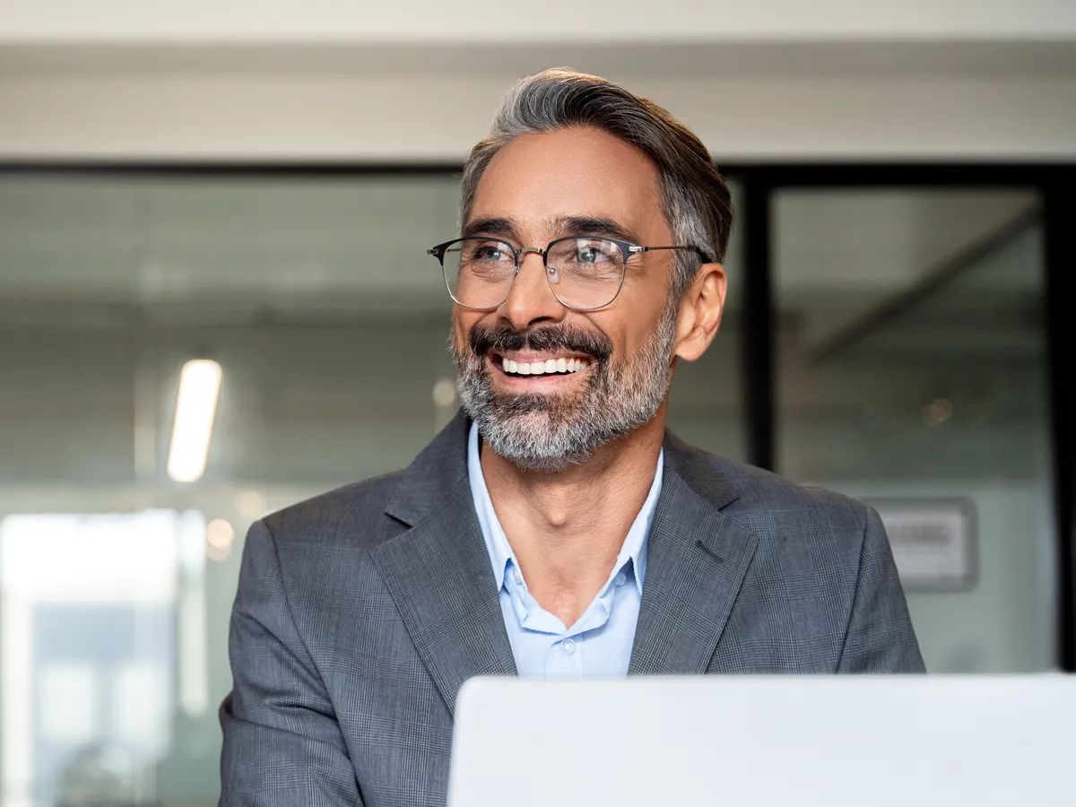 Portrait of mature Indian or Latin business man ceo trader using laptop computer, typing, working in modern office. Middle-age Hispanic smiling handsome businessman entrepreneur looking aside,dreaming