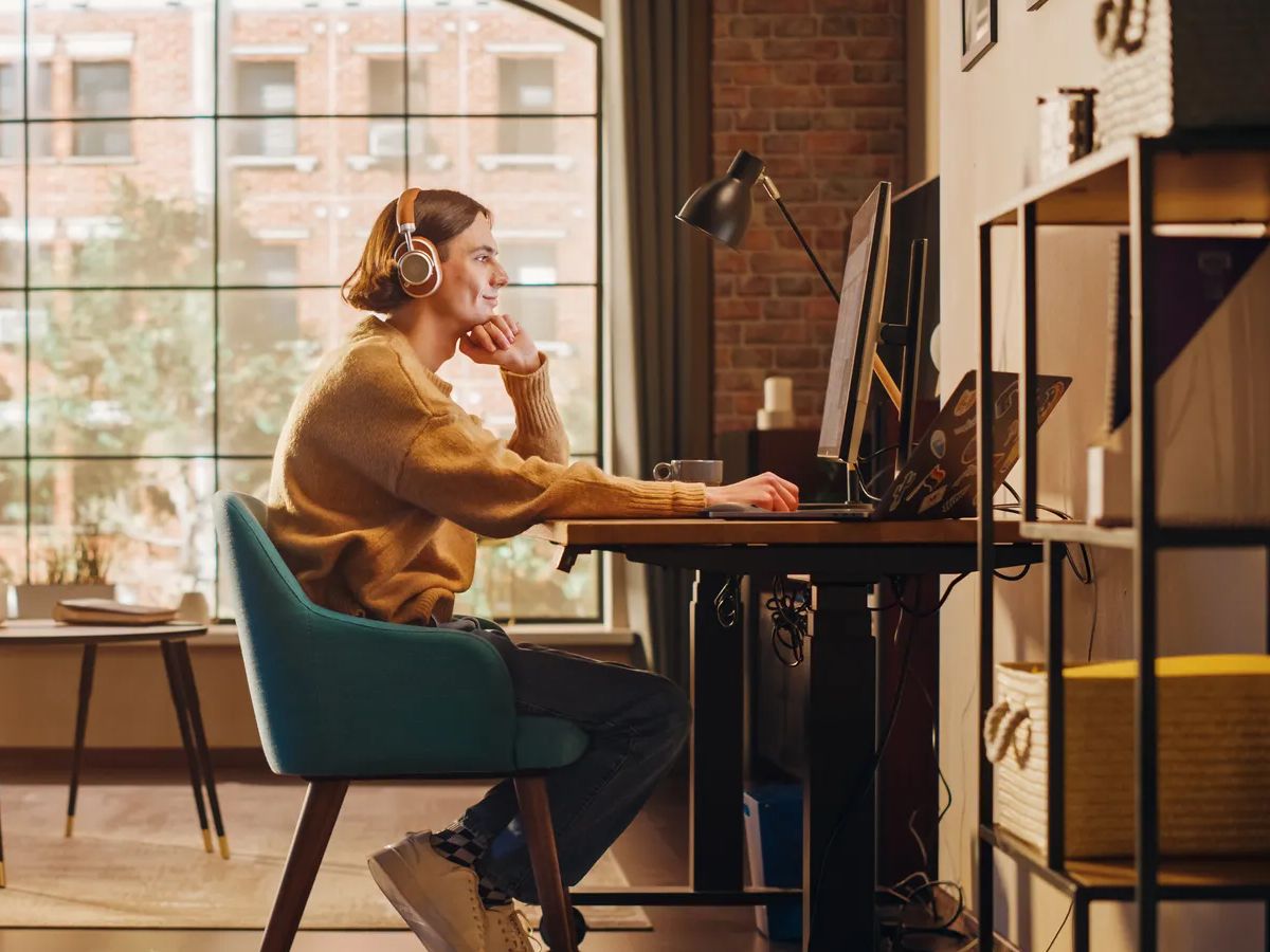 Young Handsome Man Working from Home on Desktop Computer in Sunny Stylish Loft Apartment. Creative Designer Wearing Cozy Yellow Sweater and Headphones. Urban City View from Big Window.