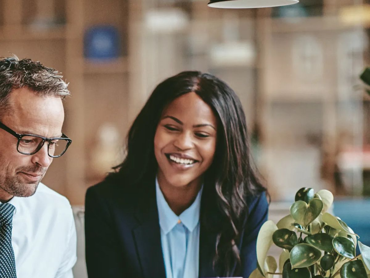 Two diverse businesspeople smiling while working on a laptop together at the end of a boardroom table in an office