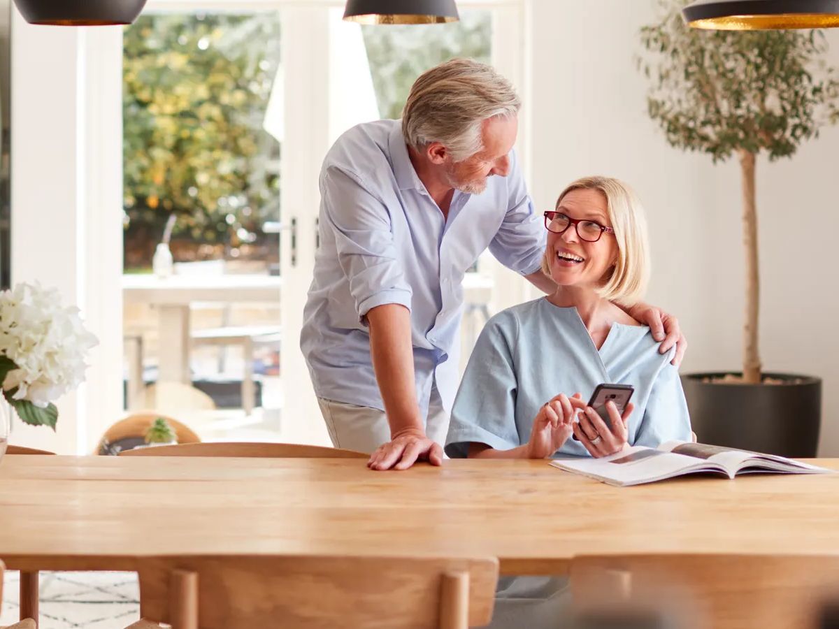 Senior Couple Relaxing With Magazine At Home Looking At Mobile Phone Sitting At Dining Room Table