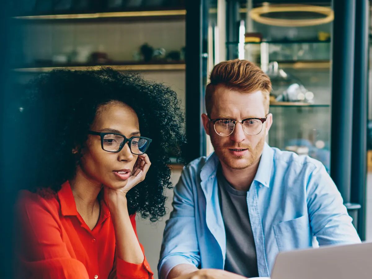 Pensive african american young woman pointing on laptop and explaining task caucasian graphic designer during meeting in coffee shop.Multicultural colleagues discussing creating website at computer