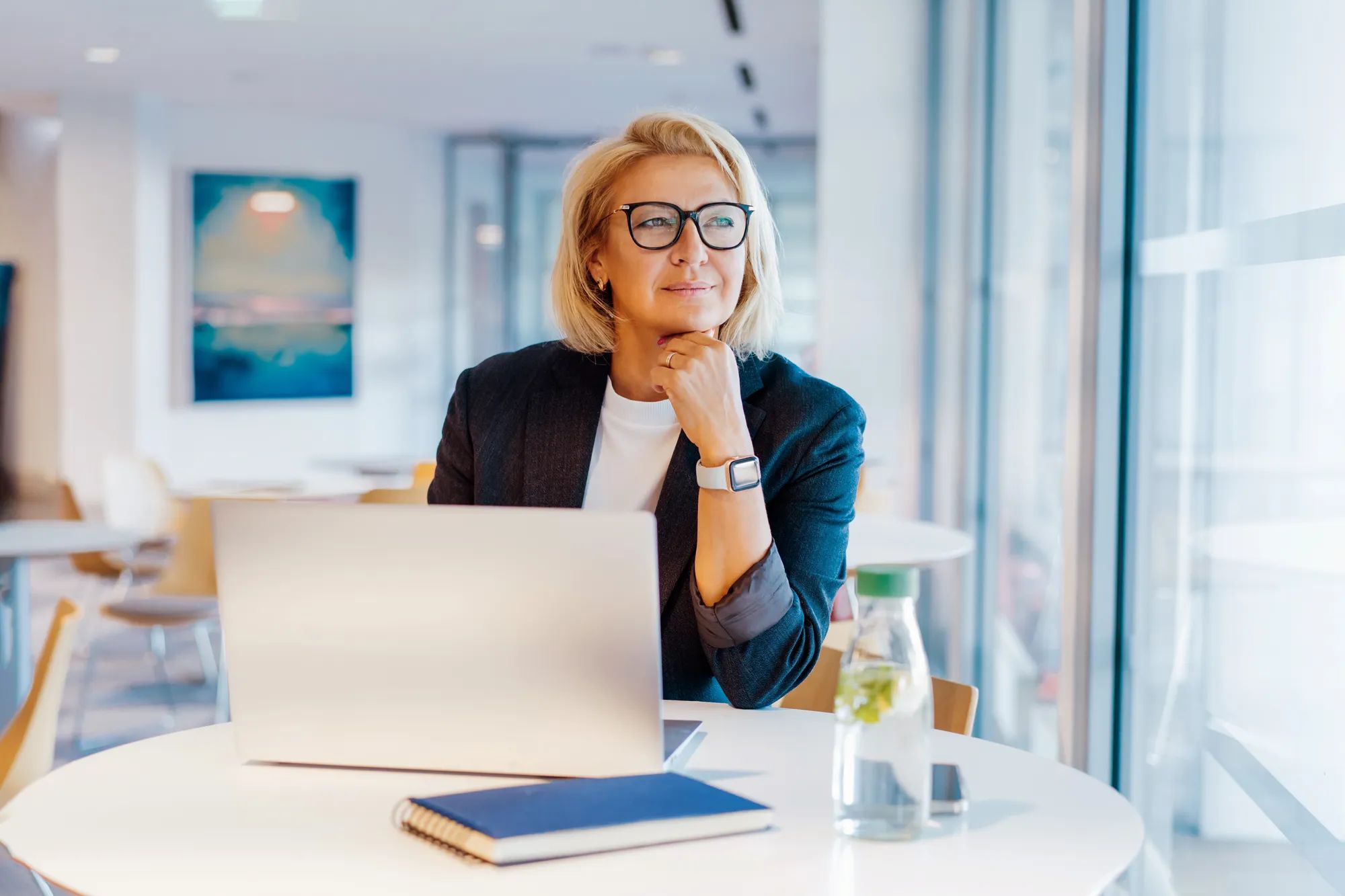 50's confident mature businesswoman dreamy looking at window, middle-aged experienced senior female professional working on laptop in open space office. Female entrepreneur working remotely