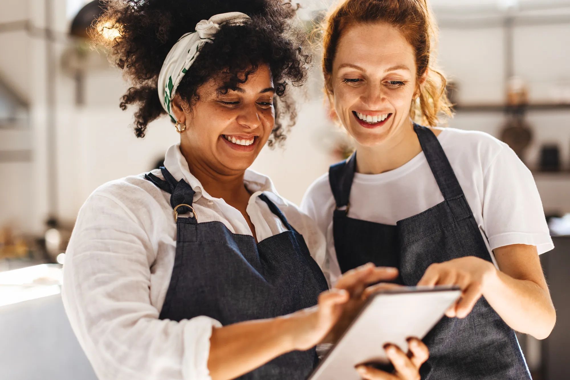 Happy restaurant workers stand side by side, focused on the touchscreen tablet in front of them. Two waitresses managing orders in their cafe, ensuring every customer receives their desired meal.