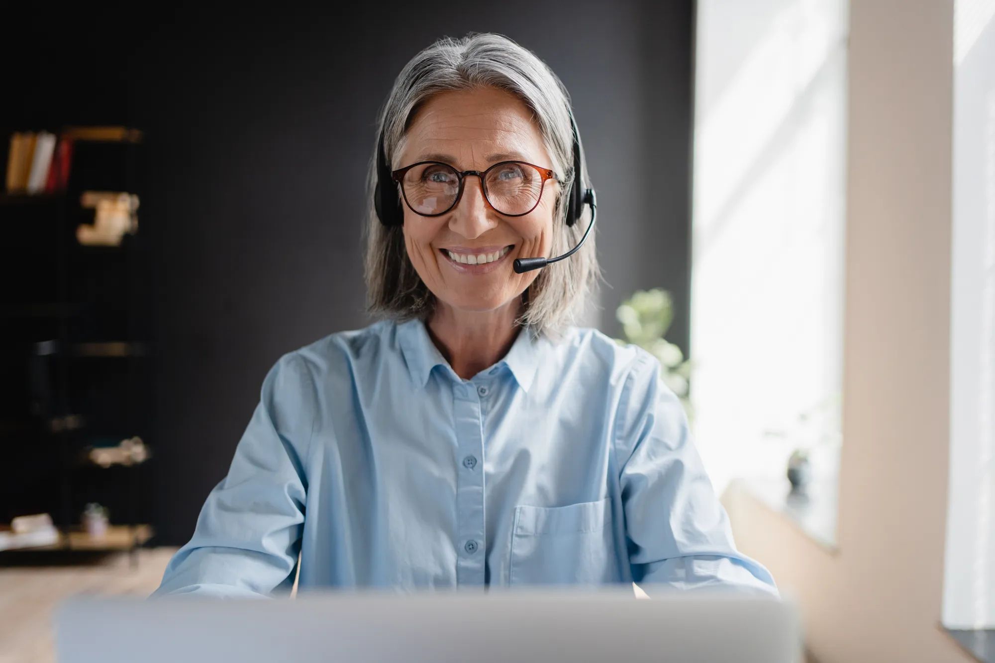 Busy mature business woman call center representative customer support agent helping client, smiling middle aged senior female operator wearing headset working using laptop computer in office.