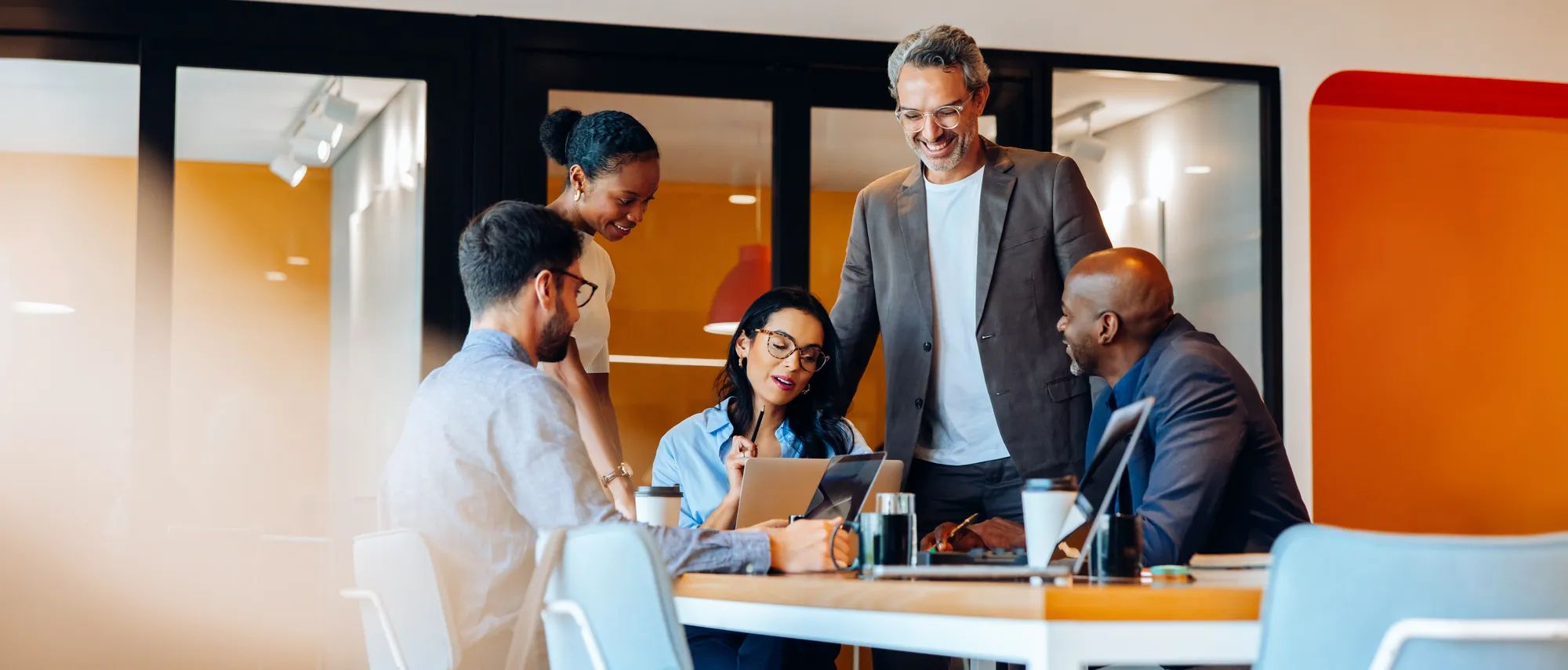 Team of five young and senior adults engaging in conversation at a workplace. They are brainstorming and utilizing technology, creating a professional yet engaging atmosphere, sitting around a table.