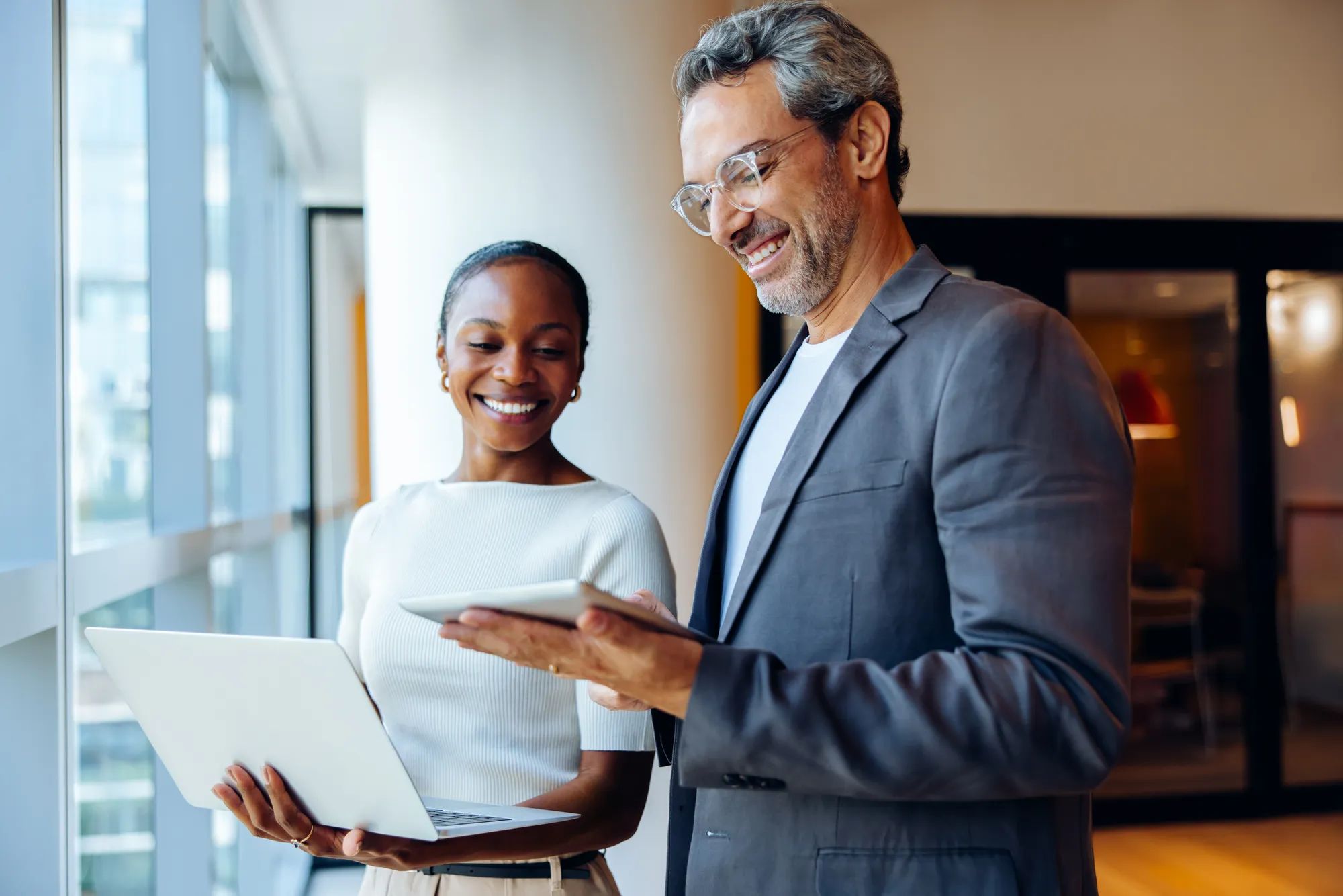 Two smiling business colleagues engaging in discussion while holding a laptop and tablet indoors. They appear in high spirits as they interact in a brightly lit setting.