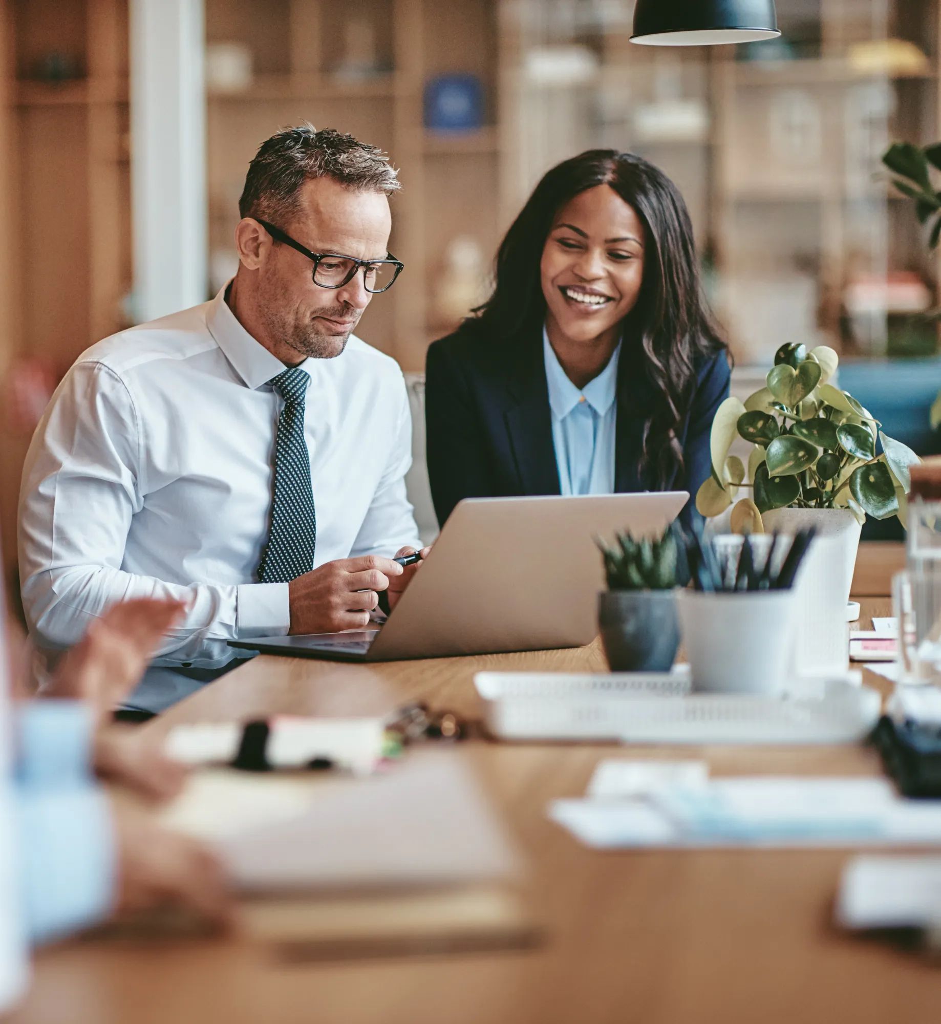 Two diverse businesspeople smiling while working on a laptop together at the end of a boardroom table in an office