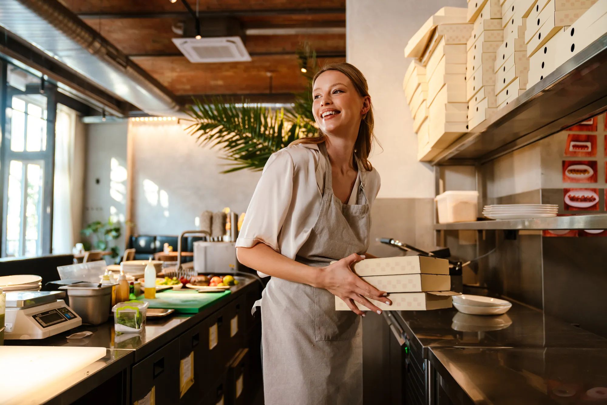 Young white woman wearing apron holding food boxes while working in restaurant kitchen