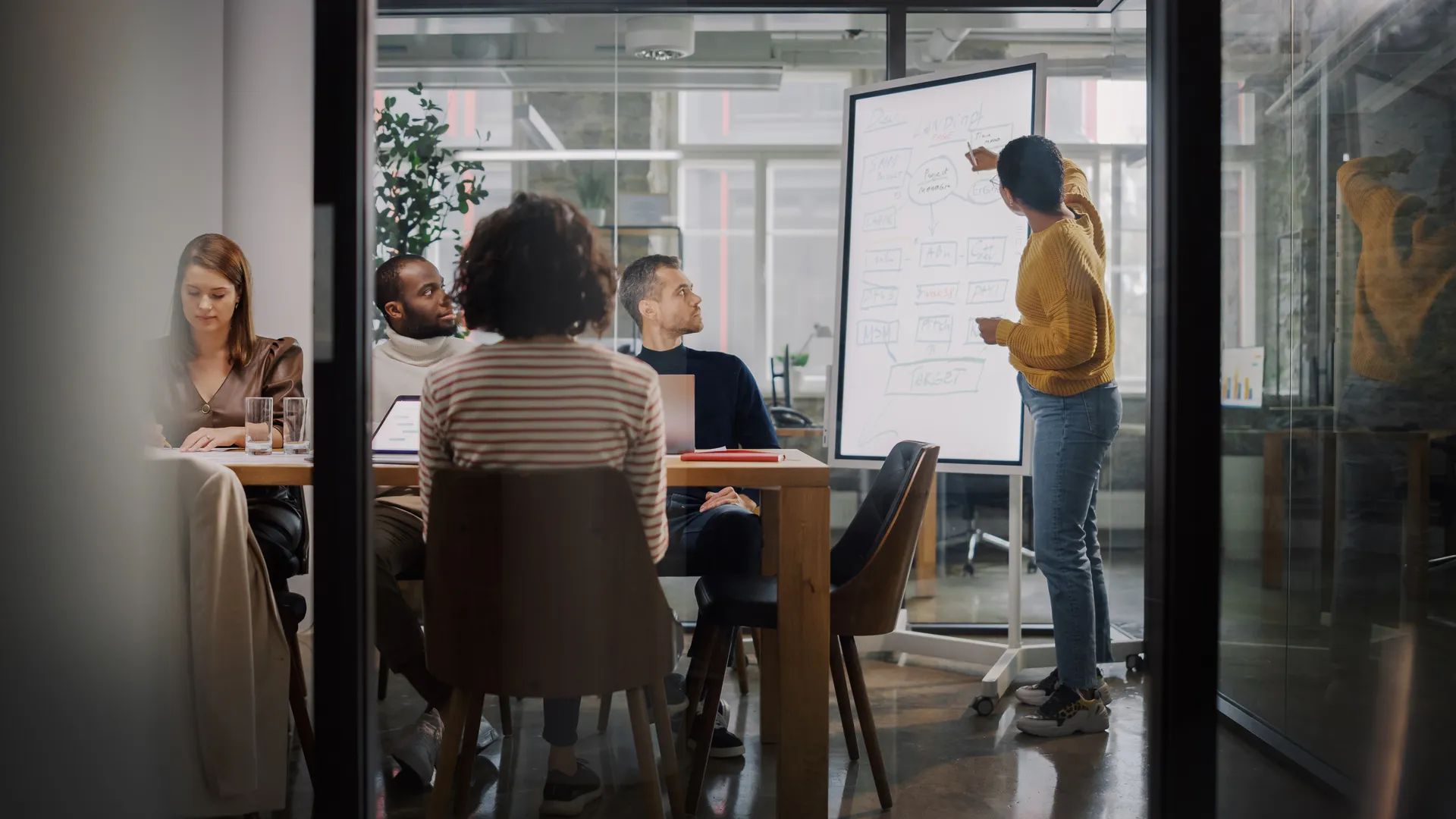 Project Manager Makes a Presentation for a Young Diverse Creative Team in Meeting Room in an Agency. Colleagues Sit Behind Conference Table and Discuss Business Development, User Interface and Design.