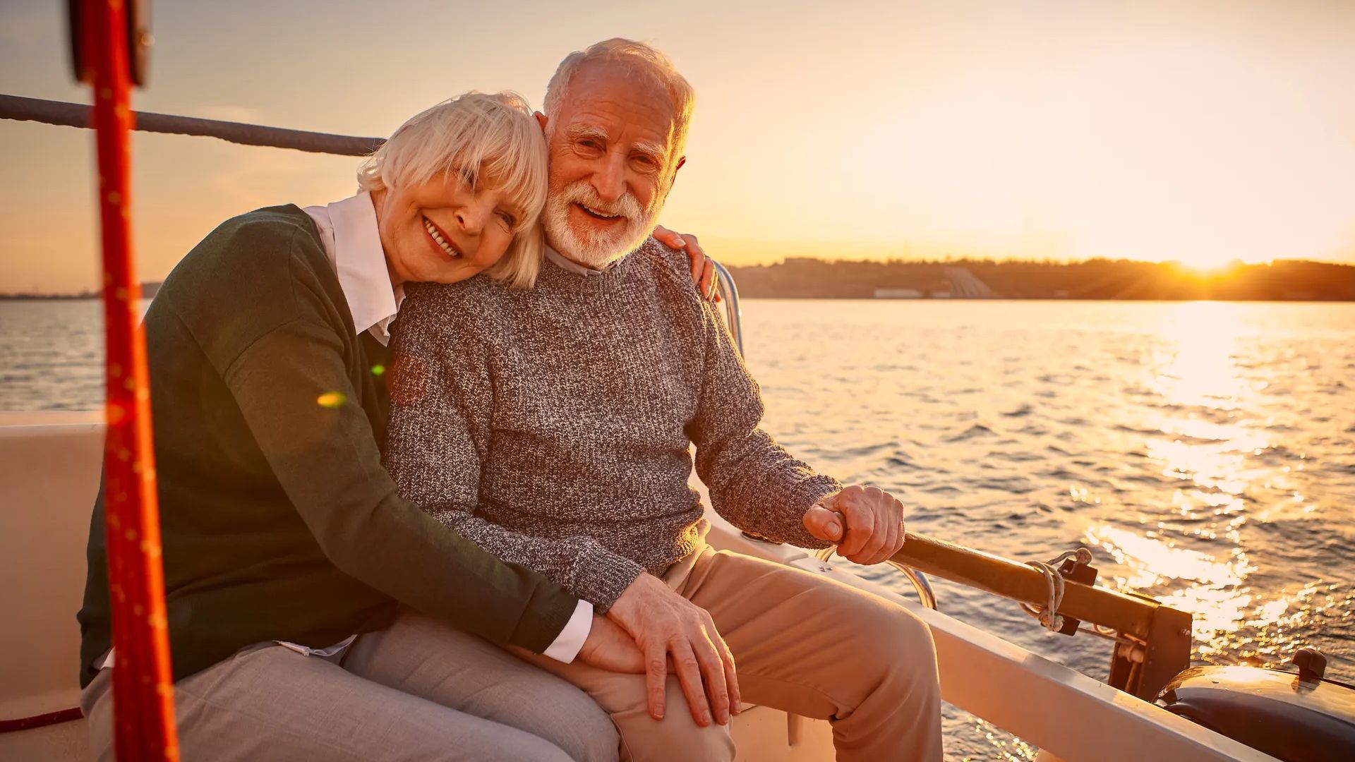 Enjoying amazing sunset. Happy senior couple, elderly man and woman holding hands, hugging and relaxing together while sitting on the side of yacht deck floating in the sea. Romantic vacation