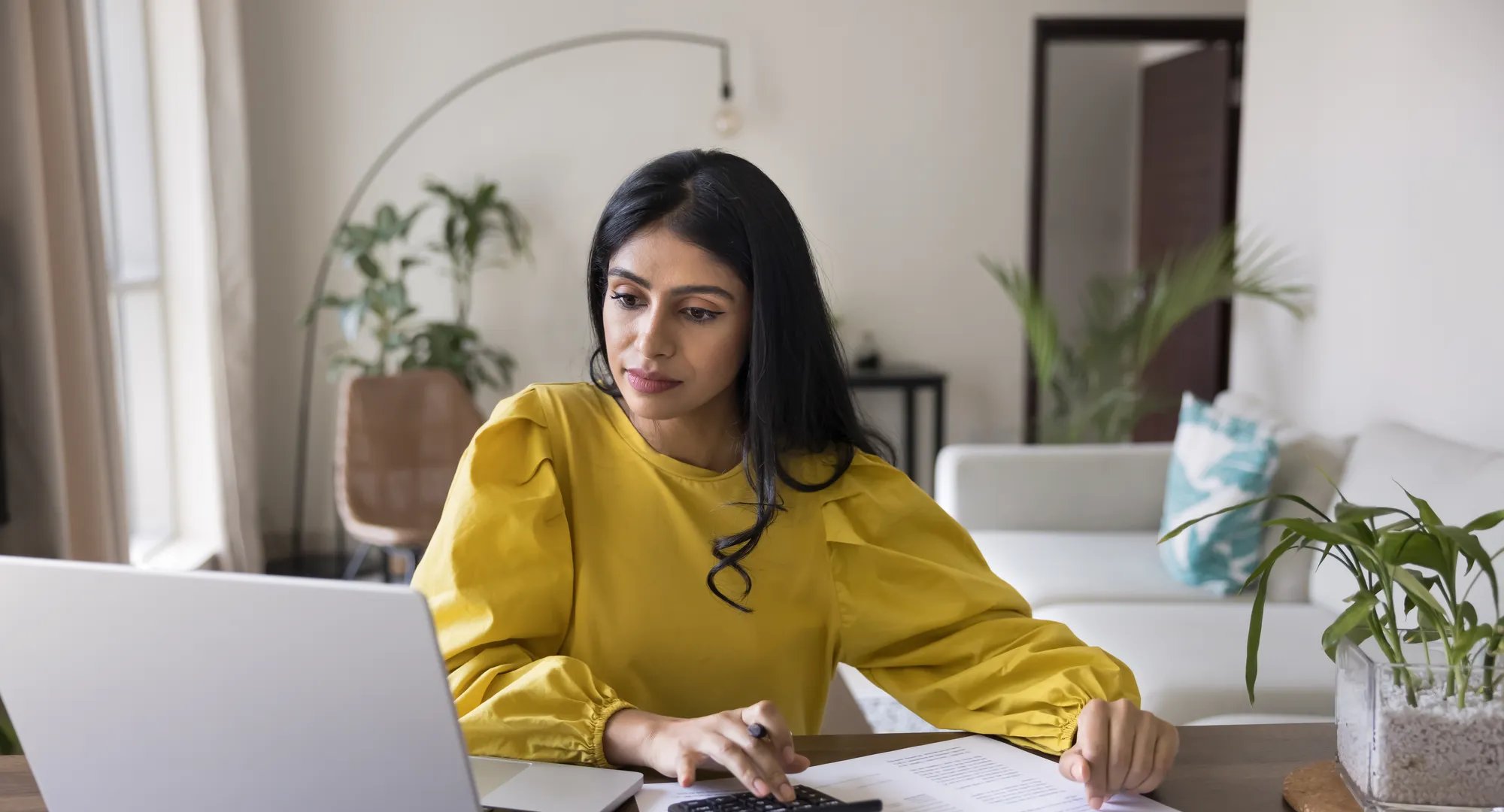 Young Indian woman sit at table with papers and laptop, controlling personal incomes and monthly expenses, reviewing invoices or bills to pay, calculating and preparing tax returns, managing finances