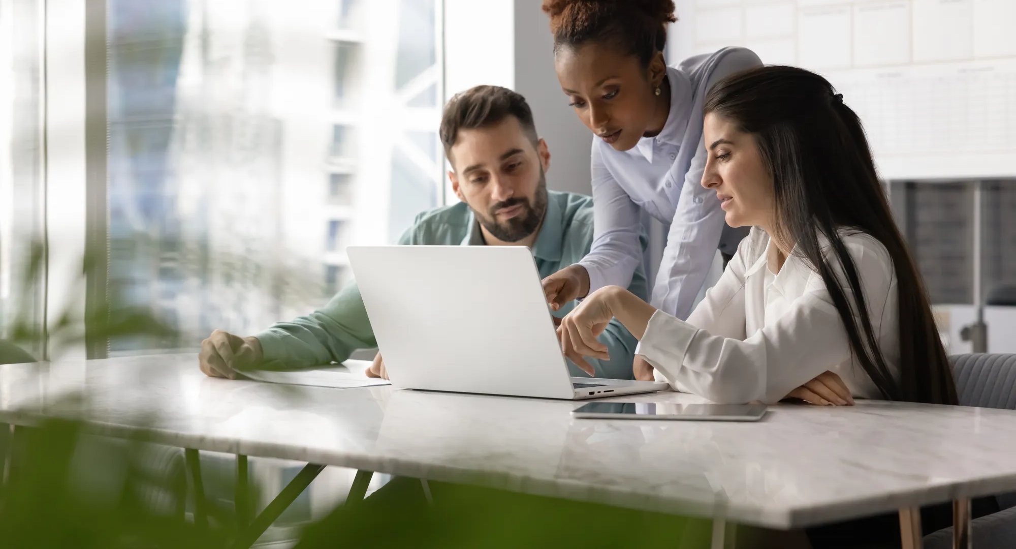 Multiethnic group of office colleagues discussing Internet marketing strategy, using laptop for teamwork, meeting at computer in office collaboration room. Candid view with border of blurred plants