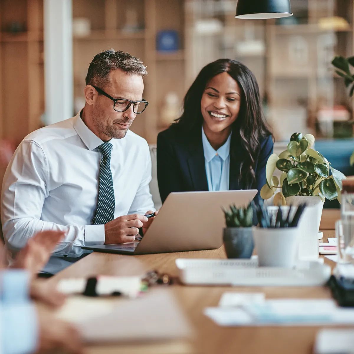Two diverse businesspeople smiling while working on a laptop together at the end of a boardroom table in an office