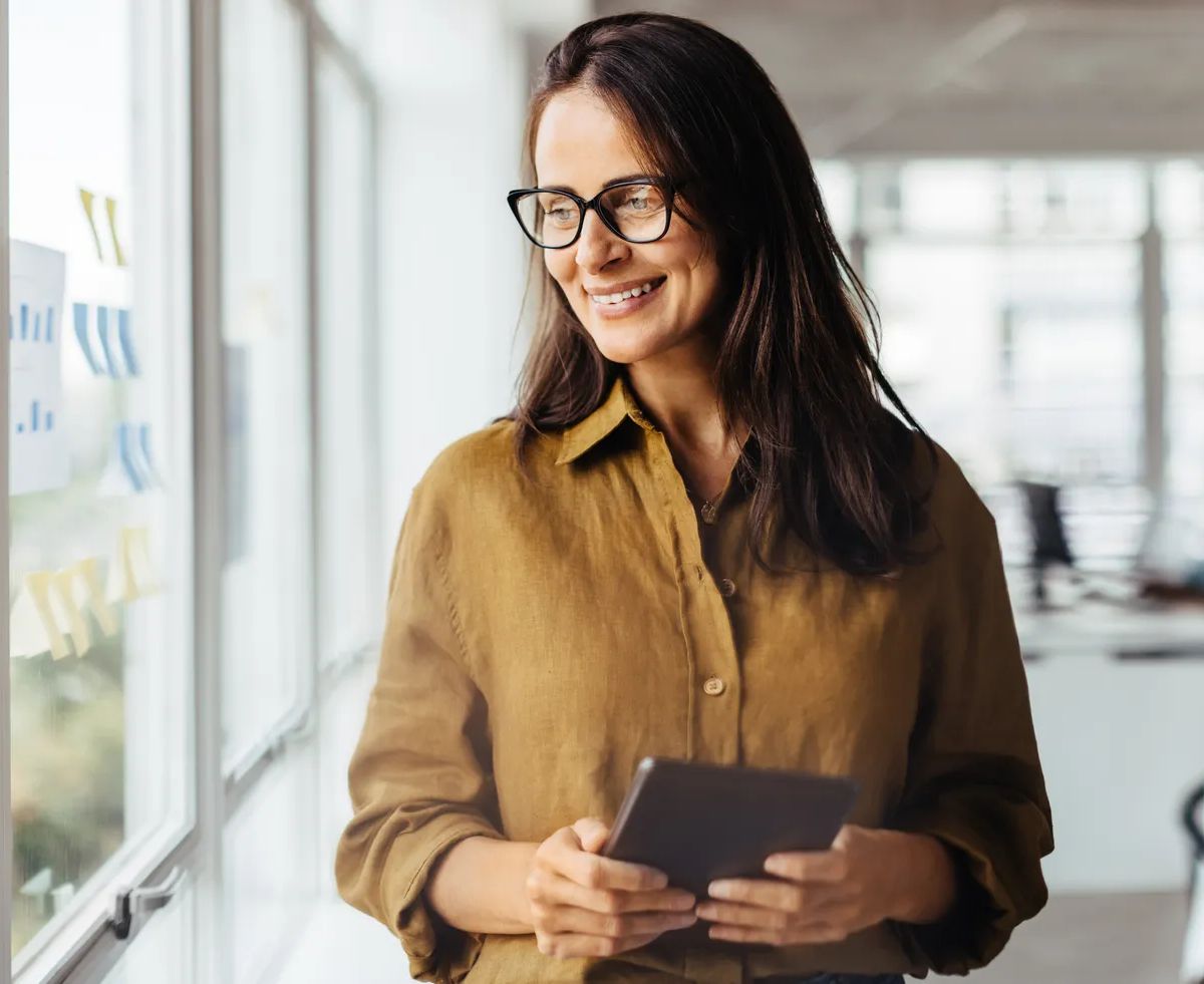 Business woman thinking while holding a tablet pc. Mature sales executive standing in an office with her colleagues in the background.