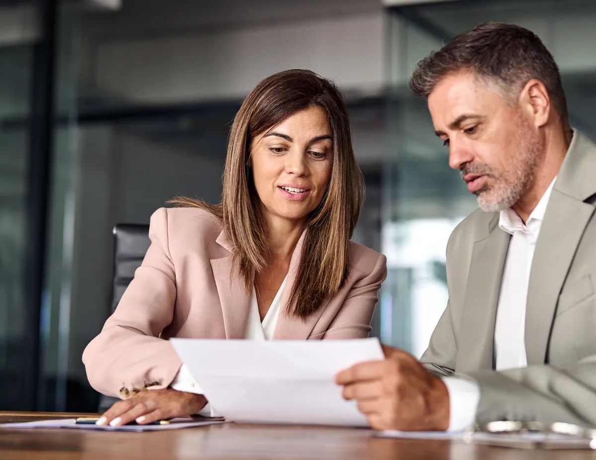 Two professional executives discussing financial accounting papers working together in office. Mature business woman manager consulting older man client holding legal documents at meeting.