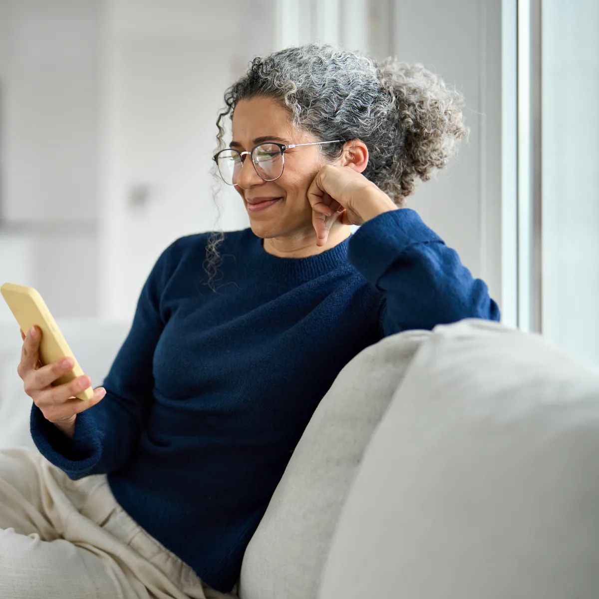Happy middle aged older woman using smartphone sitting on couch at home. Smiling mature lady holding cellphone browsing internet, texting messages, doing online shopping on mobile cell phone on sofa.