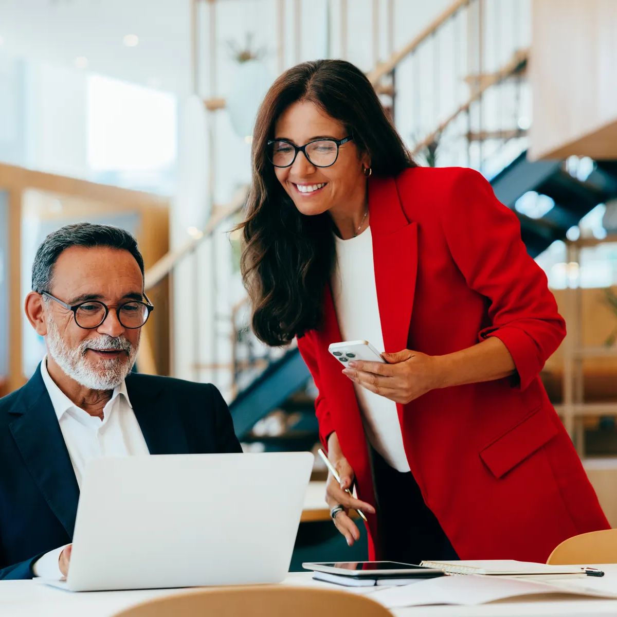 An older businessman and a younger businesswoman discussing work while reviewing a laptop screen in an office.