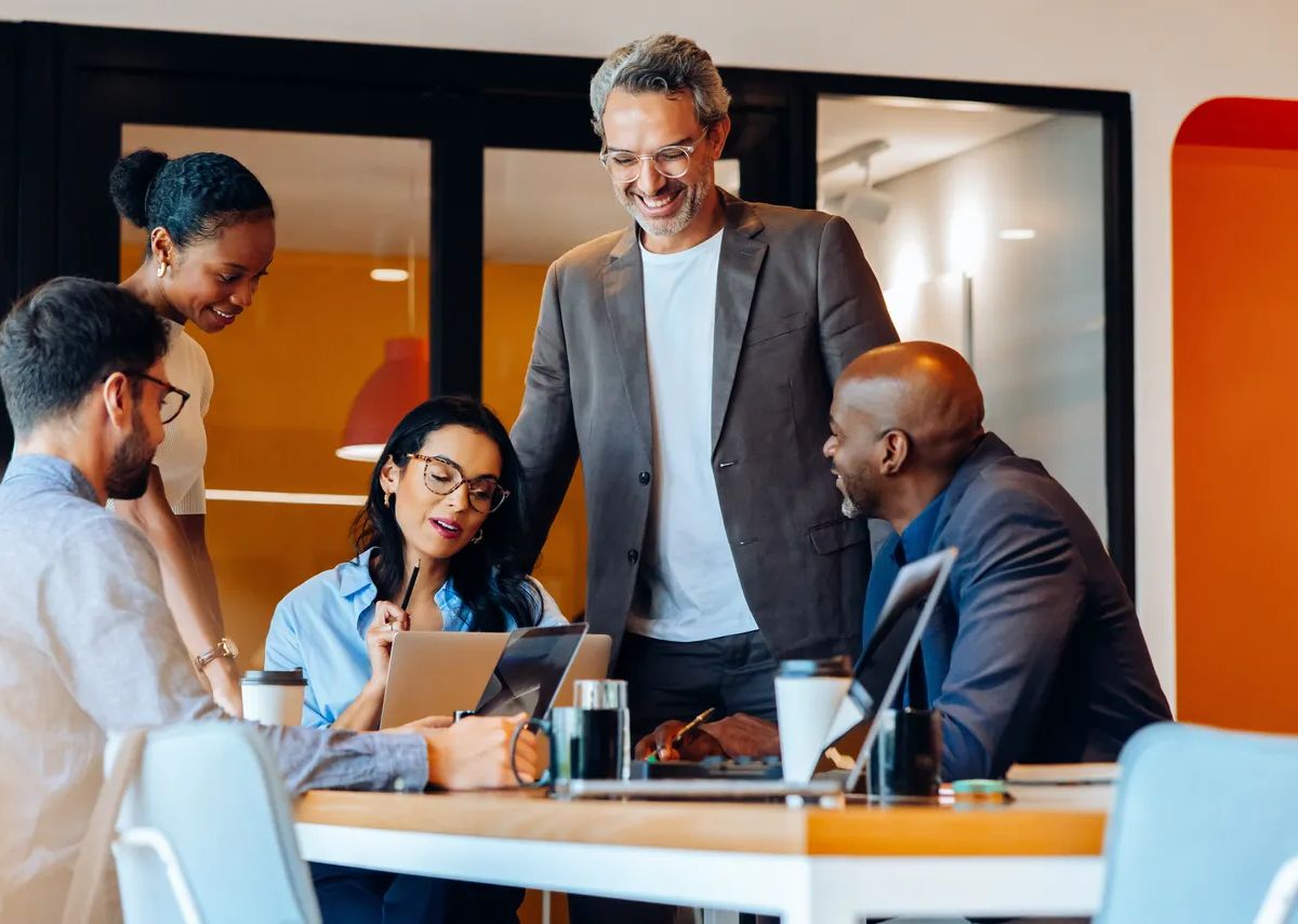 Team of five young and senior adults engaging in conversation at a workplace. They are brainstorming and utilizing technology, creating a professional yet engaging atmosphere, sitting around a table.