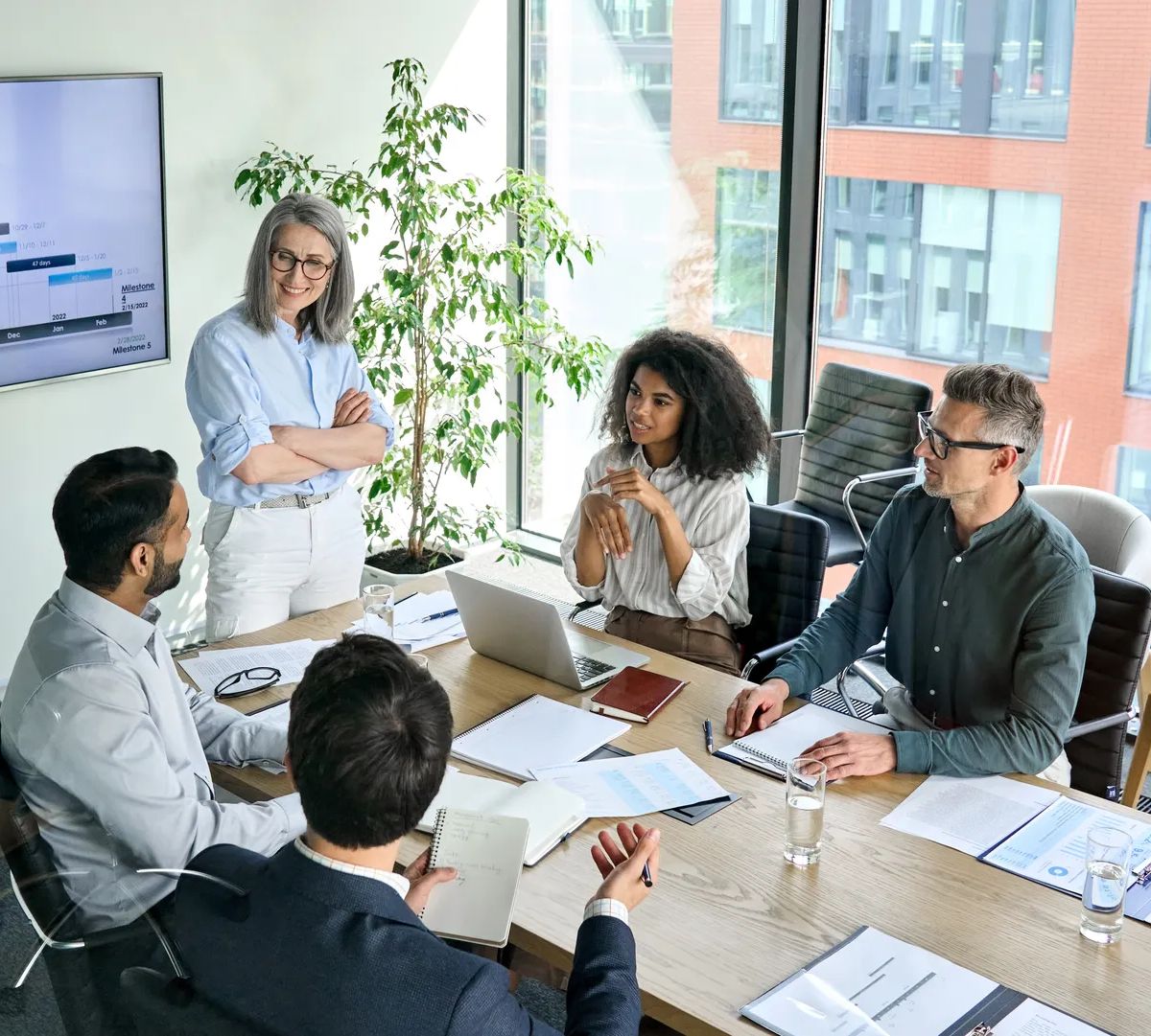 Senior female ceo and multicultural business people discussing company presentation at boardroom table. Diverse corporate team working together in modern meeting room office. Top view through glass