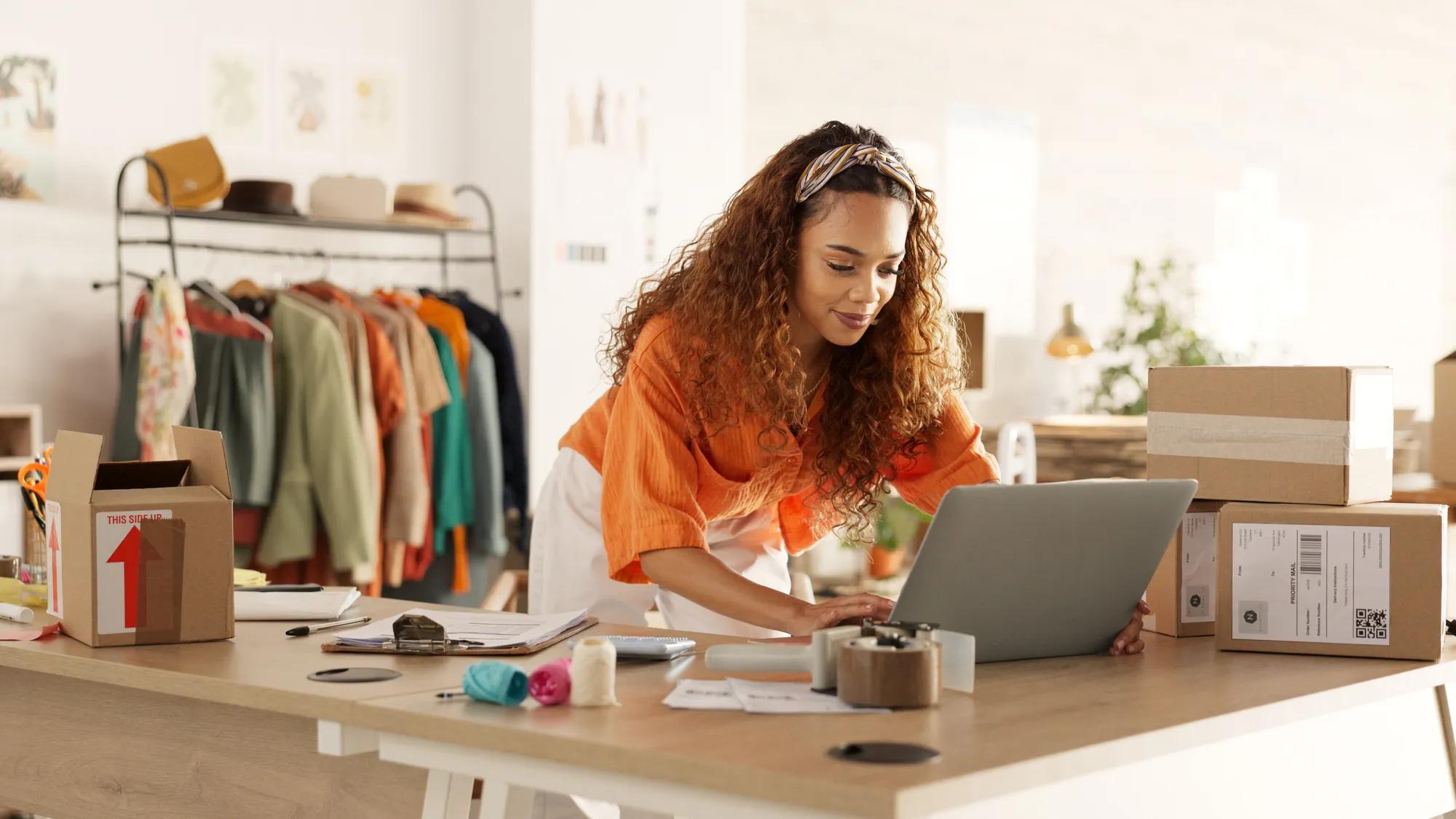 Logistics, laptop and fashion manager of retail or boutique shop checking clothing stock in boxes on a checklist. Small business, entrepreneur and young woman working with clothes inventory in store.