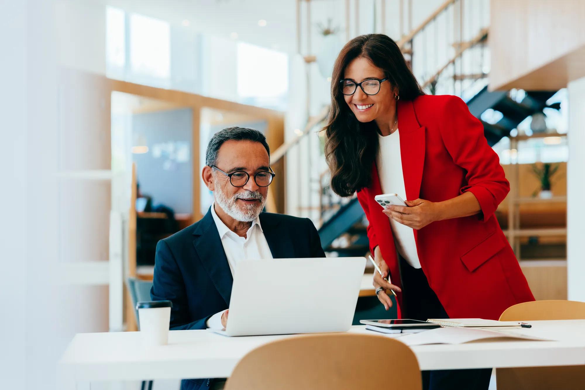 An older businessman and a younger businesswoman discussing work while reviewing a laptop screen in an office.